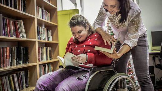 A teenage girl in a wheelchair reading a book with a support person